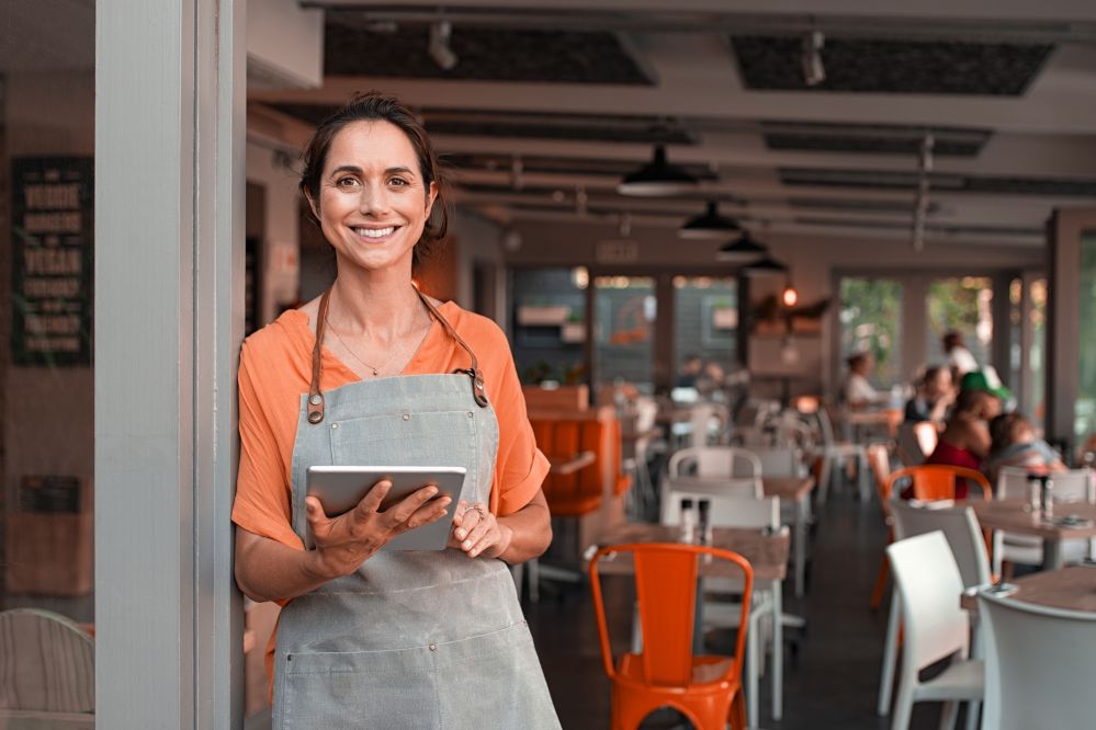 Lady standing in a doorway of a cafe business, she is holsding a ipad smiling wearing a apron and orange t-shirt