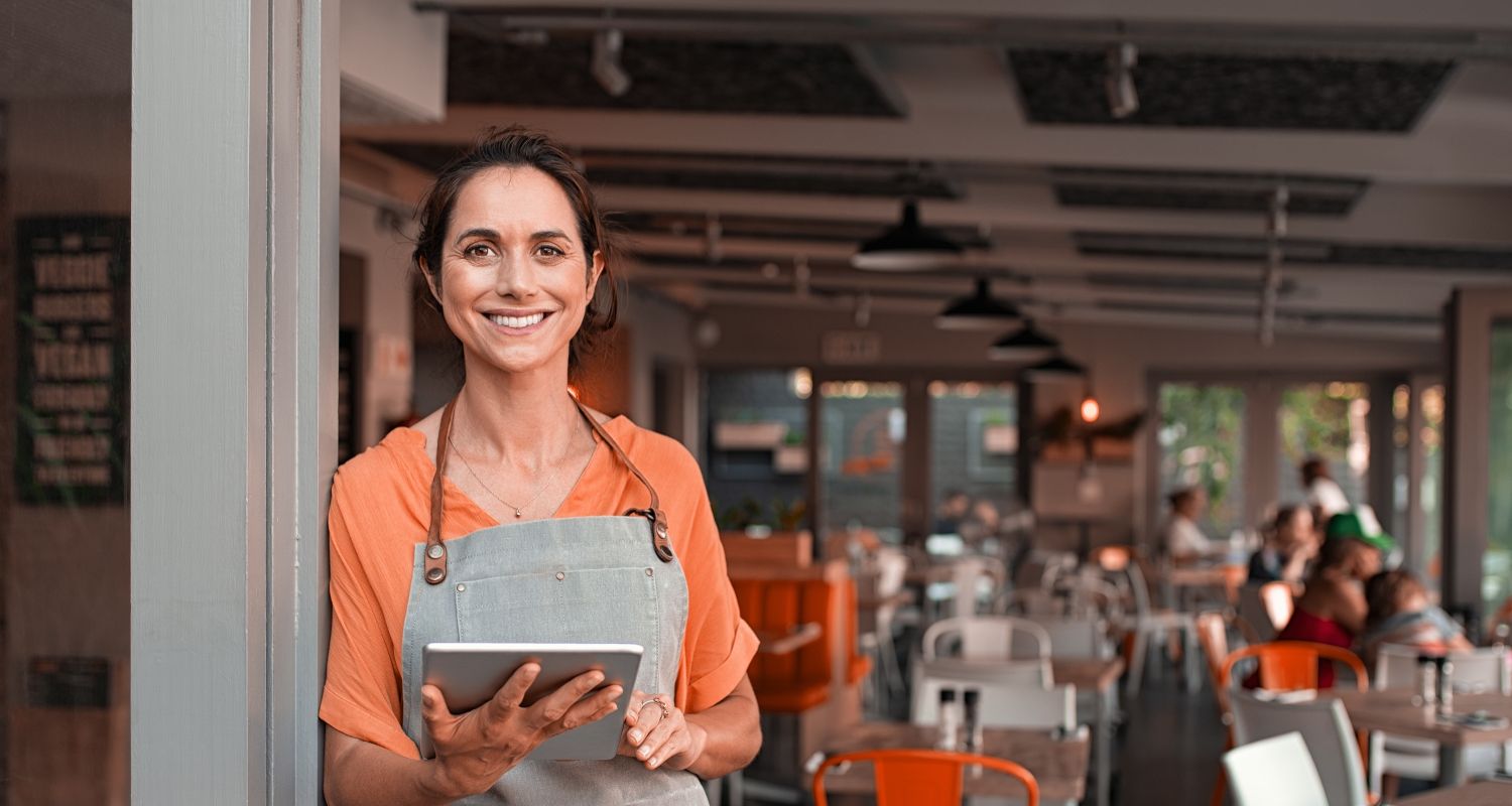 Lady standing in a doorway of a cafe business, she is holsding a ipad smiling wearing a apron and orange t-shirt